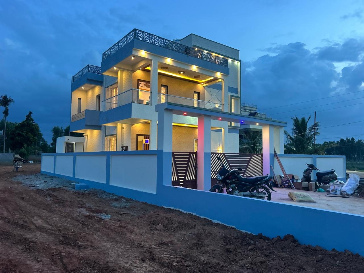 Blue-and-white 2,000 sqft duplex house in Malur at dusk, with a jaali terrace parapet, glass balcony railing and a lit car porch